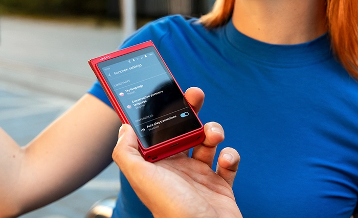 A person holds a small red electronic device displaying a language translation app, while another person in a blue shirt stands nearby.