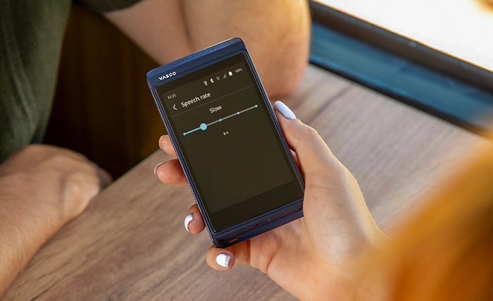 A person holds a VASCO electronic translator, displaying a screen with a Speech rate slider set to Slow. Another persons arm rests on a wooden table nearby.