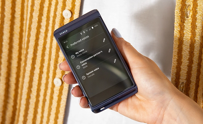 A hand holds a portable electronic device displaying a Preferred voices settings menu with options for user voice, conversation partner, and system voice. The person wears a yellow and white textured garment.